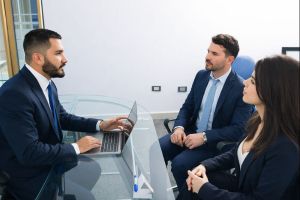 Three business professionals in a modern office meeting, with a mortgage adviser in a navy suit using a laptop at a glass desk while a suited man and woman listen across from him, using a Connect Mortgages branded blue colour palette, in a 300x200 image.