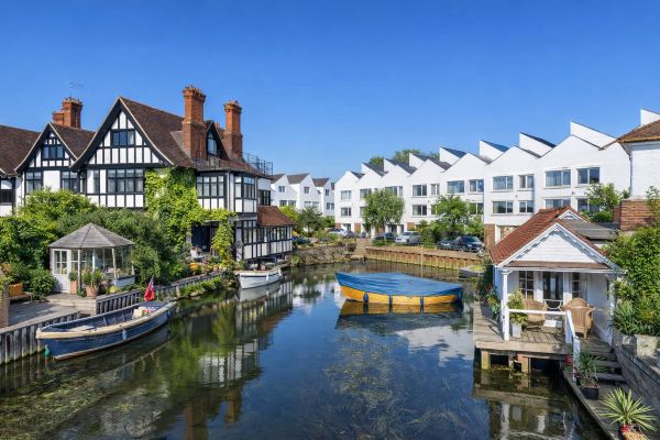 Canal-side homes with traditional Tudor-style houses and modern white townhouses reflected in calm water on a clear sunny day.