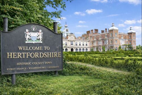 “Welcome to Hertfordshire sign set beside a historic red brick country house with formal gardens under a clear blue sky.”