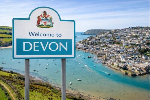 “Welcome to Devon sign overlooking a coastal harbour with turquoise water, boats, and hillside town.”