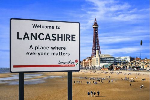 Welcome to Lancashire sign shown clean and modern in the foreground, blended with Blackpool beach and the iconic Blackpool Tower in the background, featuring a sandy shoreline, seaside buildings, and a bright blue sky.