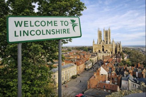 Daytime view of Lincoln in Lincolnshire showing a green “Welcome to Lincolnshire” roadside sign in the foreground, with leafy trees beside it and Lincoln Cathedral rising above historic red brick buildings and streets in the background under a clear sky. Welcome to Lincolnshire