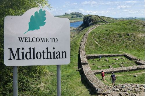 “Welcome to Midlothian roadside sign in the foreground, set against rolling green hills and historic stone ruins, with walkers following a winding path through the countryside under a bright blue sky.”