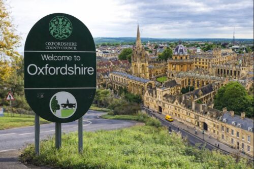 A daytime scene showing a green “Welcome to Oxfordshire” roadside sign beside a winding country road, with the historic skyline of Oxford in the background, including the Radcliffe Camera, university buildings, and church spires surrounded by trees and greenery under a bright, partly cloudy sky.