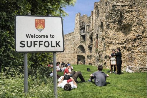 “Welcome to Suffolk roadside sign with the county crest in the foreground, set beside greenery, blended with a historic stone castle ruin in the background where people are sitting on grass and listening to a talk under a bright daytime sky.”