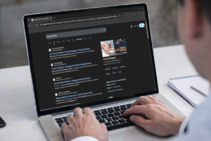 Person using a laptop on a desk while viewing Google search results for mortgage networks in dark mode. The screen shows listings for Connect Network and Connect Mortgages, along with a business profile panel displaying company details, reviews, address, and contact information. A notepad and pen sit beside the laptop, and the user’s hands are positioned on the keyboard as they browse.