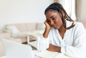 Woman sitting at a table at home, looking thoughtfully at her laptop with a notebook open, representing the question “Do I Need a Financial Adviser?”