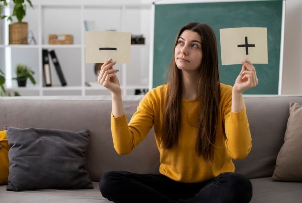 Woman sitting on a sofa holding two cards, one with a plus sign and one with a minus sign, looking thoughtful as she compares options and weighs a decision at home.