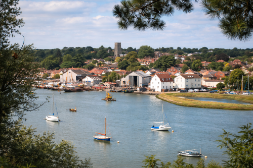 Scenic view of a riverside town with calm water in the foreground and several small boats moored on the river. Along the shoreline are white and red-roofed buildings, boatyards, and houses, with a church tower rising above the town in the background. Trees and greenery surround the settlement, and overhanging branches frame the scene under a lightly clouded sky.