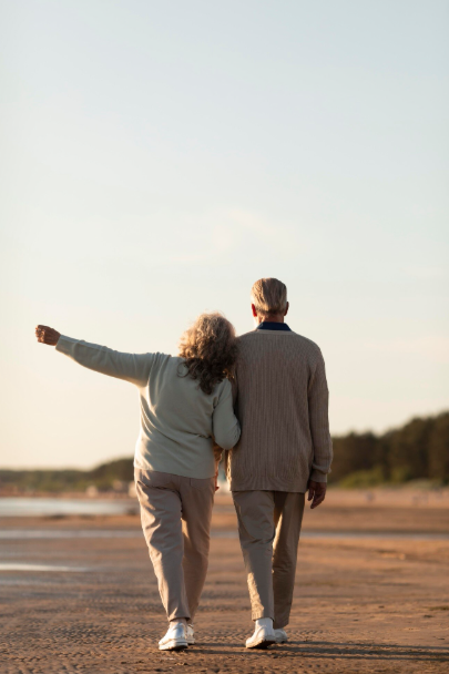 An older couple walking arm in arm along a sandy beach at sunset, viewed from behind. One person gestures outward while they stroll together near the shoreline under a clear sky.