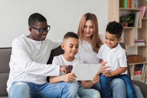 A smiling family of four sitting together on a sofa at home, looking at a tablet. The father is pointing at the screen while two young boys and their mother watch with interest, creating a warm and relaxed atmosphere.