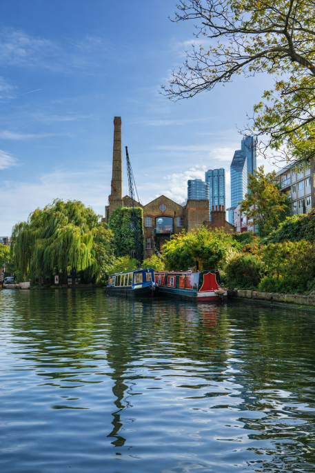 Canal boats moored along a green waterside in Islington with historic brick buildings and modern skyline in the background. PBSBrokers