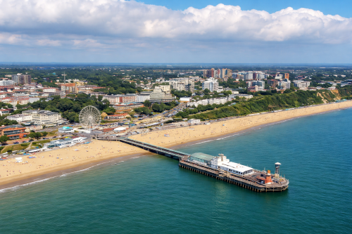 High resolution aerial view of a coastal town with a long sandy beach, turquoise sea, and a traditional pier extending into the water. The image shows a large observation wheel near the promenade, green cliffs behind the beach, and a mix of mid rise buildings and trees across the town under a partly cloudy sky.