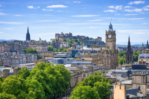 Daytime cityscape of Edinburgh showing historic stone buildings, a prominent clock tower, and a hilltop castle in the background. The image includes green trees in the foreground and a clear blue sky with light clouds above the skyline.