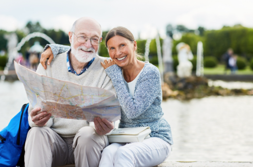 An older couple sitting by a fountain in a park smile at the camera while holding and looking at a city map together. The man has white hair and a beard and wears glasses and a light jumper, while the woman, dressed in a blue cardigan and white trousers, has her arm around his shoulders.
