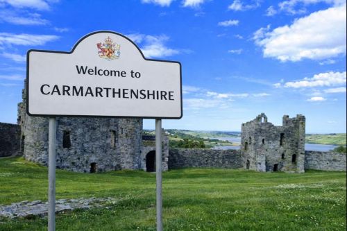 Welcome to Carmarthenshire sign with county crest, set in front of historic stone castle ruins and green countryside under a blue sky.