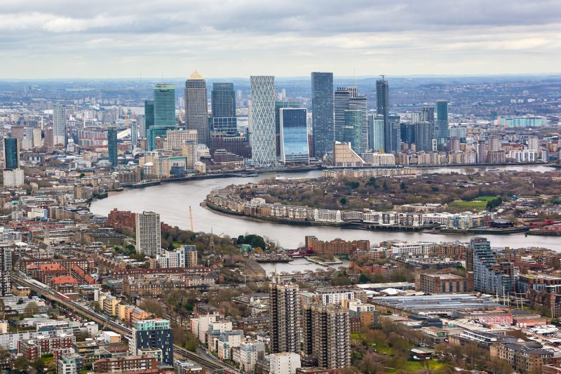 Aerial view of London’s Canary Wharf skyline with modern skyscrapers clustered along a bend in the River Thames, surrounded by dense residential neighbourhoods under a cloudy sky.