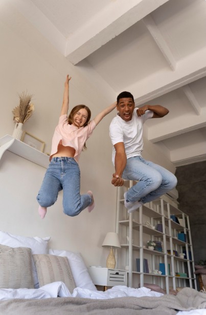 Young couple jumping joyfully on a bed in a bright, modern bedroom, celebrating together in a new home.