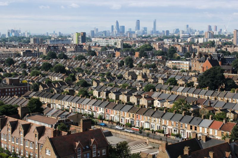 Aerial view of rows of Victorian terraced houses in London, with tree-lined streets and residential rooftops in the foreground and the city skyline, including modern skyscrapers, visible in the distance.