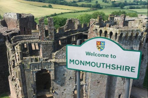 Welcome to Monmouthshire sign with county crest, positioned in front of historic castle ruins overlooking green Welsh countryside.