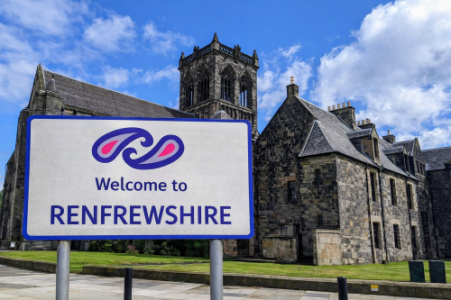 Welcome to Renfrewshire sign featuring the Renfrewshire Council logo, set in front of a historic stone abbey with a central tower under a bright blue sky with scattered clouds.