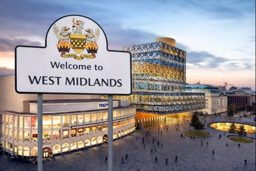 Welcome to West Midlands sign featuring the regional coat of arms, set in a city square with the Library of Birmingham and surrounding buildings at dusk.