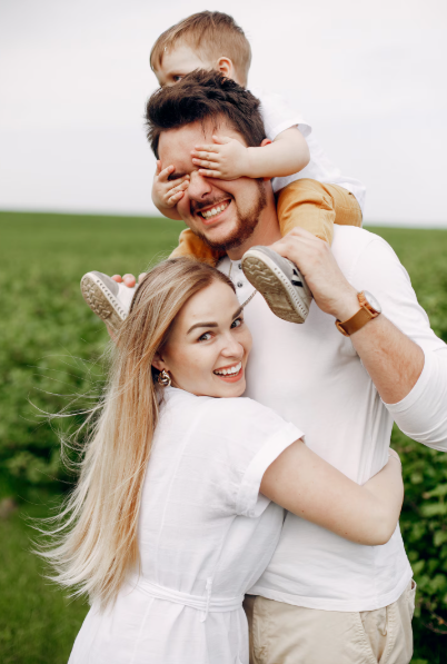 A smiling family outdoors in a green field. A man wearing a white top is carrying a young child on his shoulders, with the child playfully covering the man’s eyes. A woman in a white dress hugs the man from the side and looks towards the camera smiling. The scene appears relaxed and happy, with a natural countryside background.