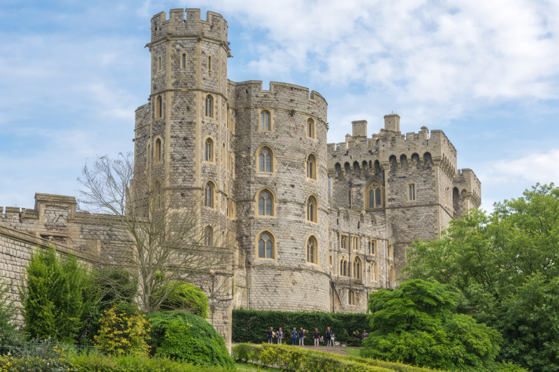 Daytime view of Windsor Castle, showing its historic stone towers and battlements surrounded by greenery under a partly cloudy sky.