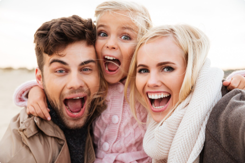 Smiling family of three taking a close-up selfie outdoors, with a mother, father, and young child laughing excitedly, conveying happiness, togetherness, and a joyful family moment.