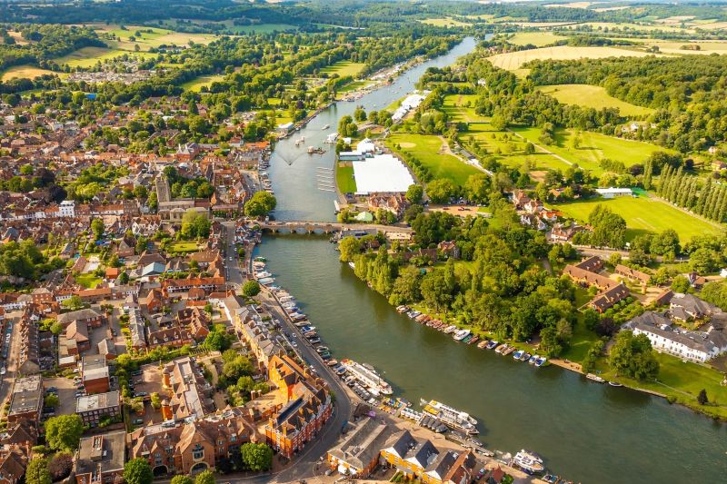 Aerial view of a riverside English town with a stone bridge crossing a winding river, surrounded by historic buildings, green parks, boats along the riverbanks, and rolling countryside in the background.