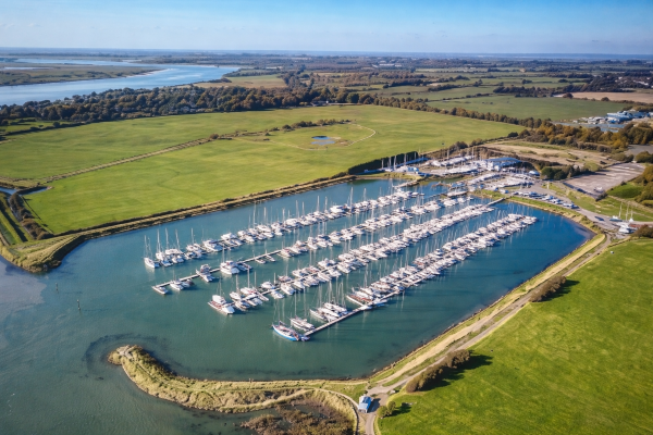 Aerial view of a marina with rows of moored boats and yachts, surrounded by green countryside and waterways under a clear blue sky.