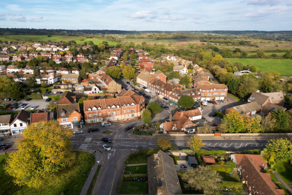 Aerial view of a small Essex town centre with rows of brick houses, shops along a main road, surrounding green fields, and countryside stretching into the distance under a partly cloudy sky.
