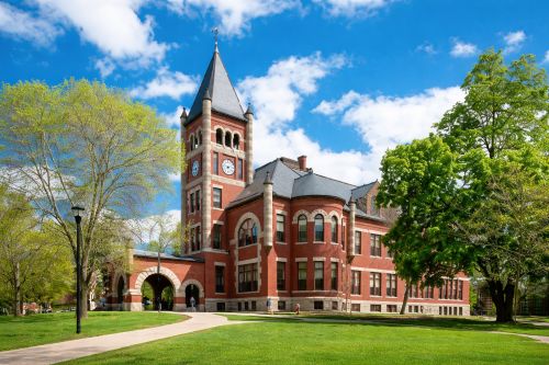 Historic red-brick civic building with a clock tower, arched entrance, and manicured lawns, surrounded by trees under a bright blue sky.