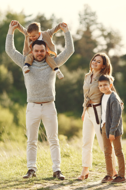 A family of four standing outdoors in a sunny natural setting. A father wearing a light sweater and trousers carries a young child on his shoulders, holding the child’s hands up playfully. Beside them, a smiling mother in light-coloured clothing stands with an older child wearing a cardigan and trousers. They are surrounded by greenery and soft sunlight, creating a warm, relaxed atmosphere.
