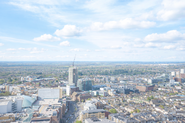 A bright, clear aerial view of a city with a mix of modern high rise buildings, residential streets, and commercial areas. A tall building under construction stands near the centre, with cranes visible. The city gradually gives way to tree lined neighbourhoods and open countryside in the distance under a blue sky with scattered white clouds.