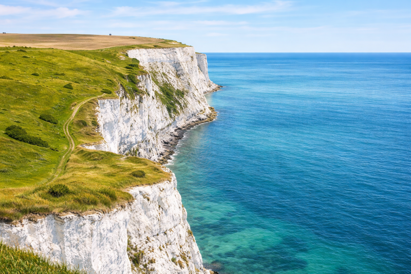 Coastal landscape with white chalk cliffs topped by green grass, overlooking a calm blue sea under a clear sky.
