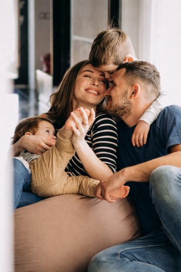 A happy family of four sitting closely together at home, with two parents smiling and holding their young children in a warm, affectionate moment.