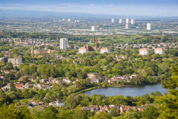 A wide aerial view of a town surrounded by greenery, showing residential neighbourhoods with houses and apartment blocks set among trees. A lake or reservoir sits in the foreground, with historic buildings and tall chimneys visible among the rooftops. In the distance, larger urban buildings rise against a backdrop of rolling hills under a clear sky.