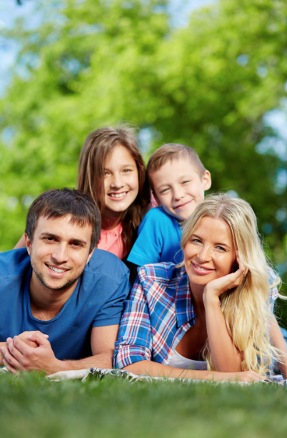 A family of four lying on the grass outdoors, smiling at the camera. Two adults are at the front with two children positioned behind them. The scene is set in a green park or garden with trees in the background, creating a relaxed and happy family atmosphere in natural surroundings.