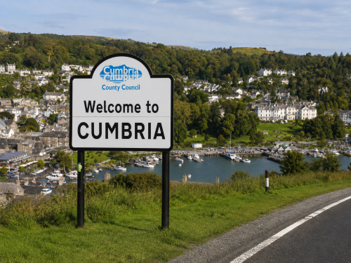 Realistic branded illustration of life in Cumbria showing a lakeside market town, green hills, mountain scenery, village homes, sailing boats, sheep, dry stone walls, and rural Lake District countryside. Why Move to Cumbria