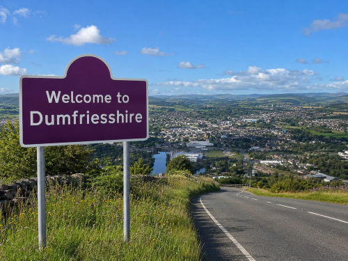 Purple “Welcome to Dumfriesshire” road sign beside a rural road, overlooking Dumfries town, green countryside, woodland and rolling hills under a blue sky. Why Move to Dumfriesshire