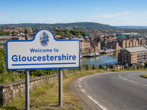 Welcome to Gloucestershire road sign beside a curved road, overlooking Gloucester Docks, city buildings, waterways and surrounding green hills under a blue sky. Why Move to Gloucestershire