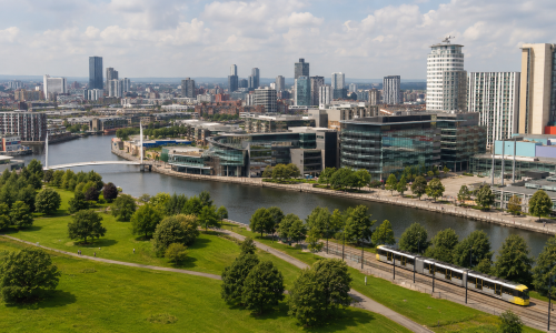 Wide view of Greater Manchester showing Manchester city centre, Salford Quays, riverside green space, modern buildings and a Metrolink tram under a cloudy sky. Why Move to Greater Manchester