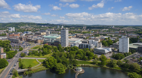 Wide aerial view of a Lanarkshire town centre with modern buildings, roads, green parkland, a pond and surrounding countryside under a bright blue sky. Why Move to Lanarkshire