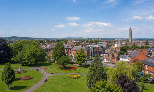 Wide view of Loughborough in Leicestershire, showing Queen’s Park, landscaped green space, red-brick town buildings and the Carillon Tower under a blue sky. Why Move to Leicestershire
