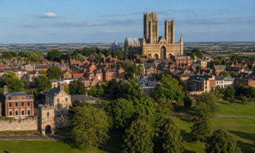 Wide aerial view of Lincoln, showing Lincoln Cathedral, historic buildings, green parkland and open countryside beyond the city. Why Move to Lincolnshire