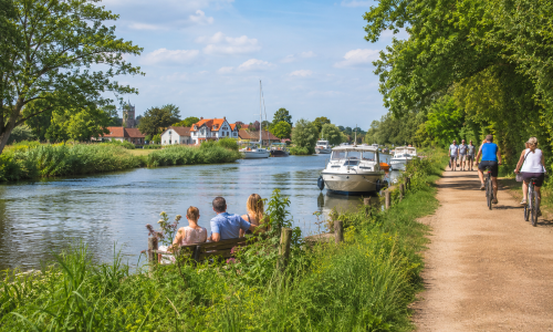 People walking, cycling and relaxing beside a Norfolk Broads waterway, with boats, reeds, village homes, trees and open sky in the background. Why Move to Norfolk
