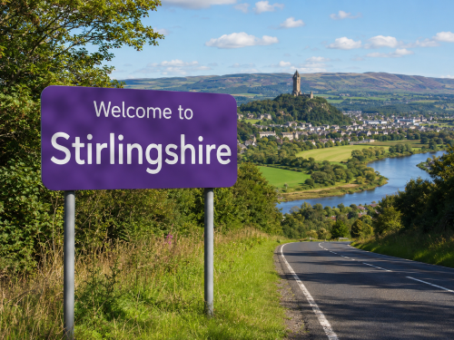 Purple “Welcome to Stirlingshire” road sign beside a country road, with green fields, a winding river, a town and a hilltop monument under a bright blue sky. Why Move to Stirlingshire