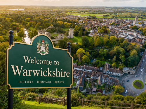 Welcome to Warwickshire road sign overlooking Warwick Castle, historic town rooftops, green parkland and countryside at sunset. Why Move to Warwickshire